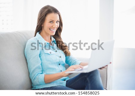 Happy brunette sitting on sofa in bright living room using laptop