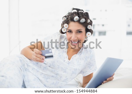 Smiling brunette in hair curlers lying on her bed using her tablet to shop online in bedroom at home