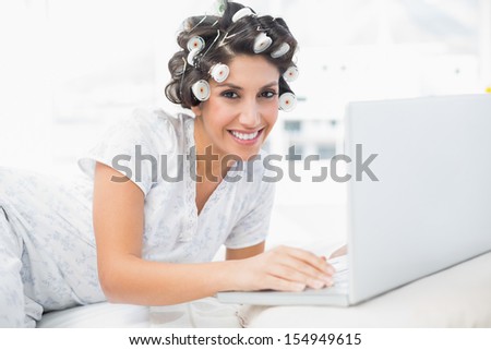 Cheerful brunette in hair rollers lying on her bed using her laptop in bedroom at home
