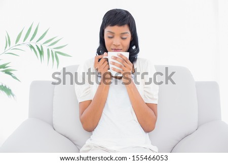 Natural black haired woman in white clothes enjoying coffee in a living room