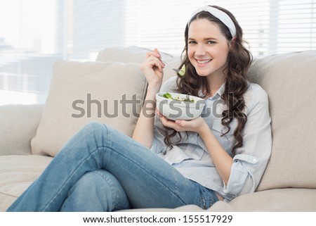 Pretty casual woman on cosy couch in bright living room eating salad