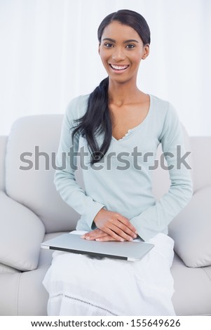 Cheerful attractive woman sitting on cosy sofa in bright living room with her laptop on her laps