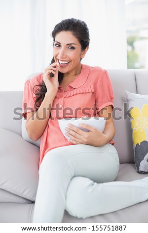 Cheerful brunette sitting on her sofa watching tv eating bowl of popcorn at home in the sitting room