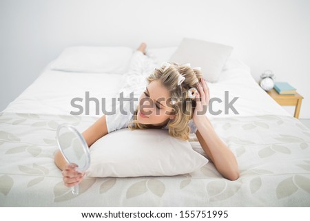 Peaceful blonde wearing hair curlers looking at reflection in mirror in bright bedroom
