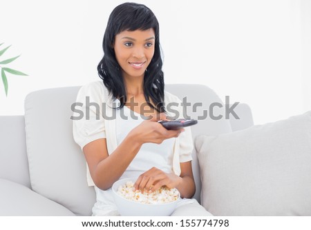Concentrated black haired woman in white clothes watching tv while eating popcorn in a living room