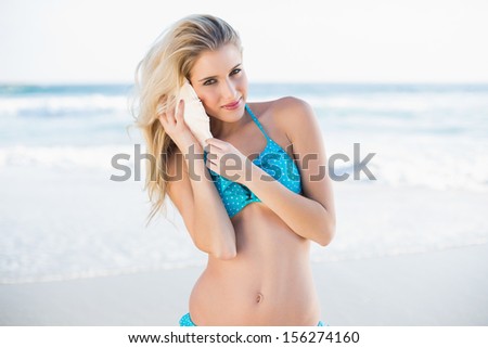Sexy gorgeous blonde in bikini holding shell by her ear on a beautiful sunny beach