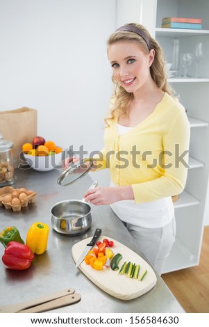 Smiling cute blonde holding cover of a pot in bright kitchen