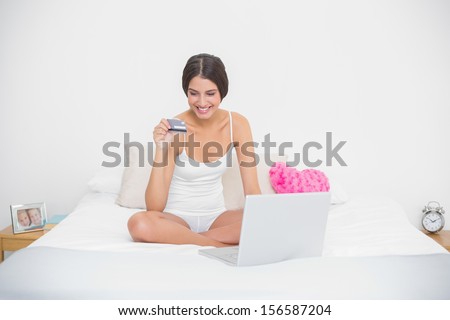 Smiling young brown haired model in white pajamas shopping online with her laptop in bright bedroom