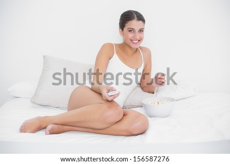Amused young brown haired model in white pajamas watching tv while eating popcorn in bright bedroom