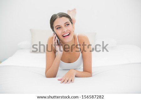 Casual young brown haired model in white pajamas calling with her mobile phone in bright bedroom