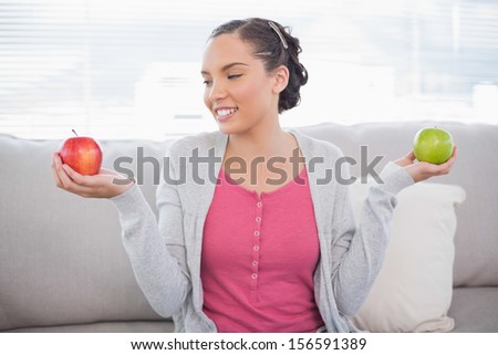 Cheerful woman sitting on sofa holding green and red apple looking at the red one in living room