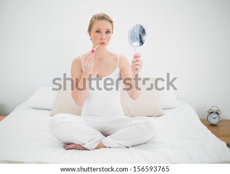 Natural stern blonde holding mirror and applying lip gloss in bright bedroom