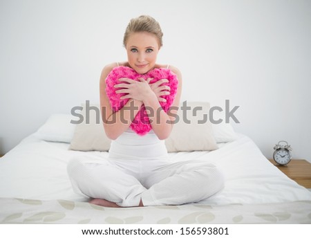 Natural content blonde holding heart pillow in bright bedroom