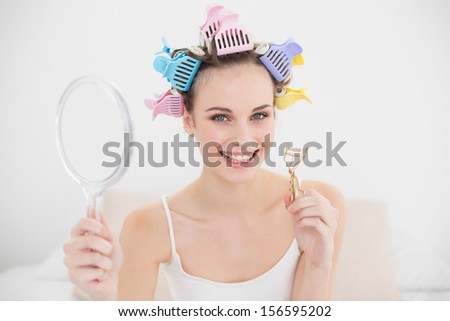 Charming natural brown haired woman in hair curlers holding a mirror and an eyelash curler in bright bedroom