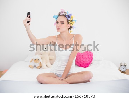 Amused natural brown haired woman in hair curlers taking a picture of herself with mobile phone in bright bedroom