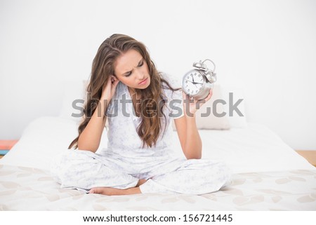 Angry brunette holding alarm clock in bright bedroom