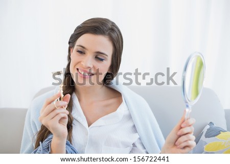Pensive casual brown haired woman in white pajamas looking at her eyelash curler in a bright living room