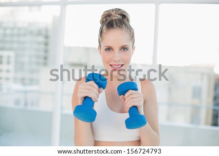 Smiling sporty blonde working out with dumbbells in bright room