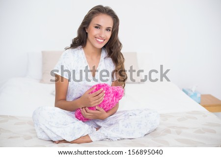 Content attractive brunette holding a heart pillow in bright bedroom