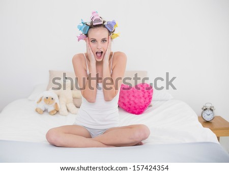 Astonished natural brown haired woman in hair curlers posing looking at camera in bright bedroom