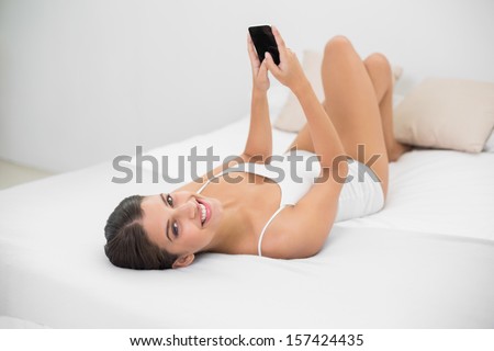 Delighted young brown haired model in white pajamas using a mobile phone in bright bedroom