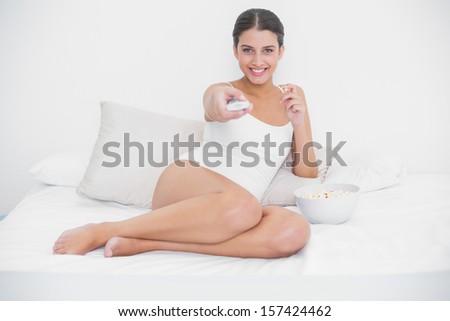 Cheerful young brown haired model in white pajamas watching tv while eating popcorn in bright bedroom