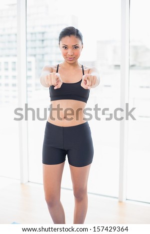 Seductive dark haired model in sportswear pointing her fingers at camera in bright fitness studio