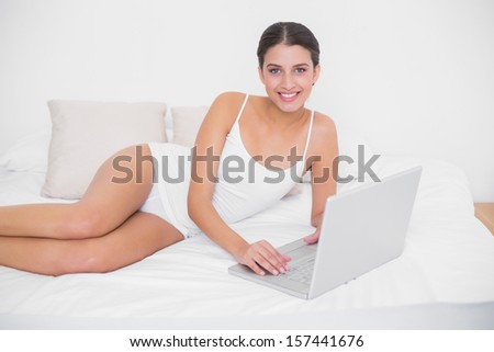 Calm young brown haired model in white pajamas using a laptop in bright bedroom
