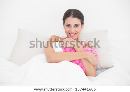 Happy young brown haired model in white pajamas hugging a heart shaped pillow in bright bedroom