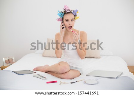 Funny natural brown haired woman in hair curlers making a phone call while applying gloss in bright bedroom
