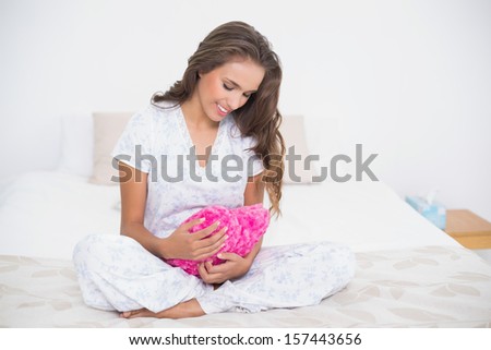 Smiling attractive brunette looking down at heart pillow in bright bedroom
