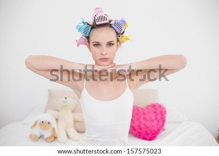 Serious natural brown haired woman in hair curlers posing looking at camera in bright bedroom