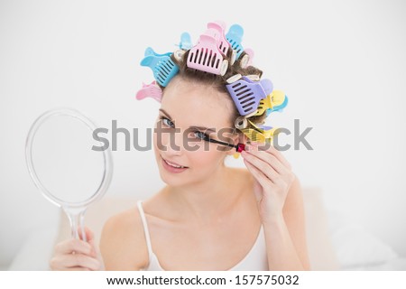 Beautiful natural brown haired woman in hair curlers applying mascara in bright bedroom