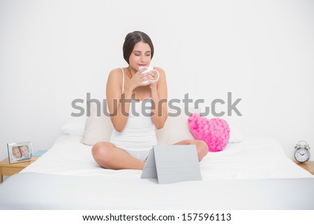 Peaceful young brown haired model in white pajamas enjoying coffee smell in bright bedroom