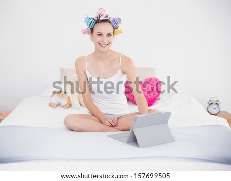 Happy natural brown haired woman in hair curlers using a tablet pc in bright bedroom