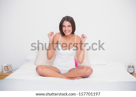 Cheerful young brown haired model in white pajamas raising her fists in bright bedroom