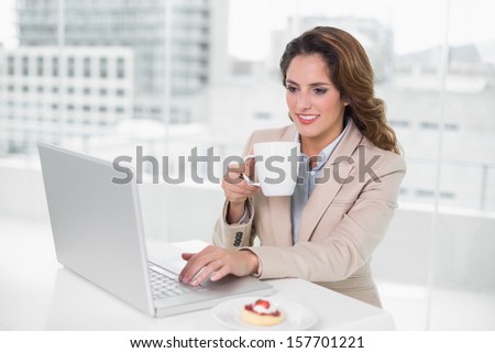 Cheerful businesswoman using laptop at her desk and holding mug in bright office