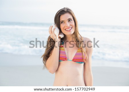 Cheerful slim brown haired model in colored bikini listening to a shell on the beach