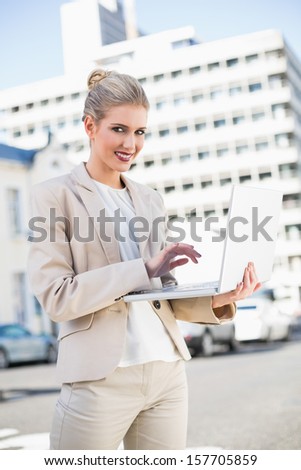 Smiling elegant businesswoman working on laptop outdoors on urban background