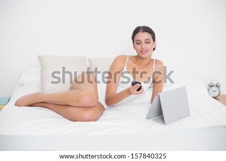 Thinking young brown haired model in white pajamas using a mobile phone in bright bedroom