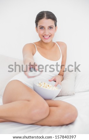 Cute young brown haired model in white pajamas eating popcorn while watching tv in bright bedroom