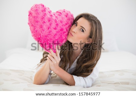 Beautiful casual brown haired woman in white pajamas kissing a heart shaped pillow in a bright bedroom