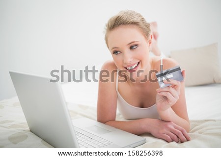Natural smiling blonde holding credit card and looking at laptop in bright bedroom