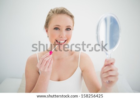 Natural smiling blonde holding mirror and applying lip gloss in bright bedroom