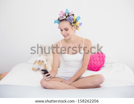 Pleased natural brown haired woman in hair curlers using her mobile phone in bright bedroom