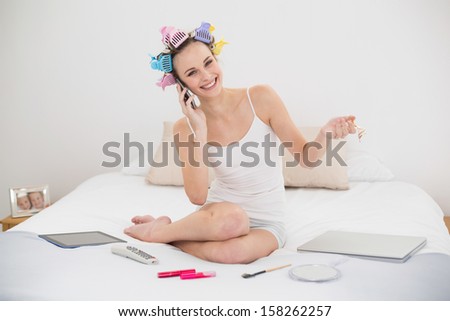 Amused natural brown haired woman in hair curlers calling with her mobile phone in bright bedroom