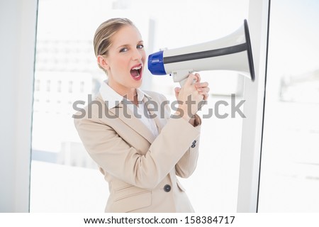 Furious blonde businesswoman in bright office shouting in megaphone