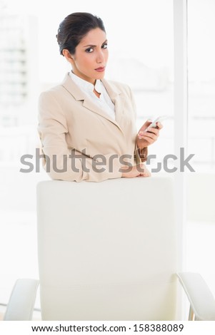 Frowning businesswoman standing behind her chair holding her phone glaring at camera in her office