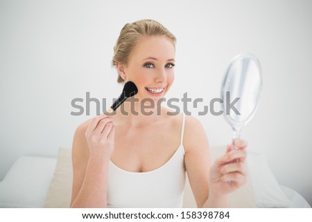Natural happy blonde holding mirror and using brush in bright bedroom