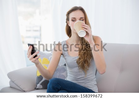 Young woman sitting on sofa drinking from disposable cup texting on the phone at home in the living room